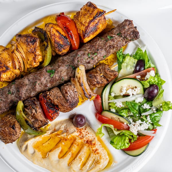 A Greek platter with chicken and beef kebabs, rice, hummus, and a fresh salad with feta.