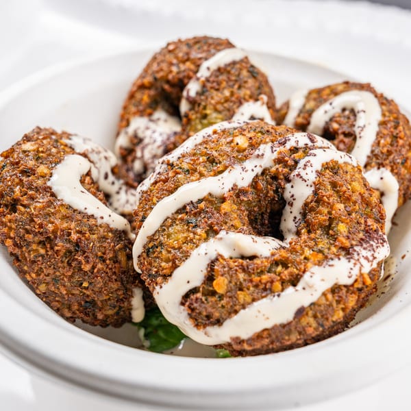 Close-up of five falafel patties drizzled with white sauce and sprinkled with spices in a white bowl.