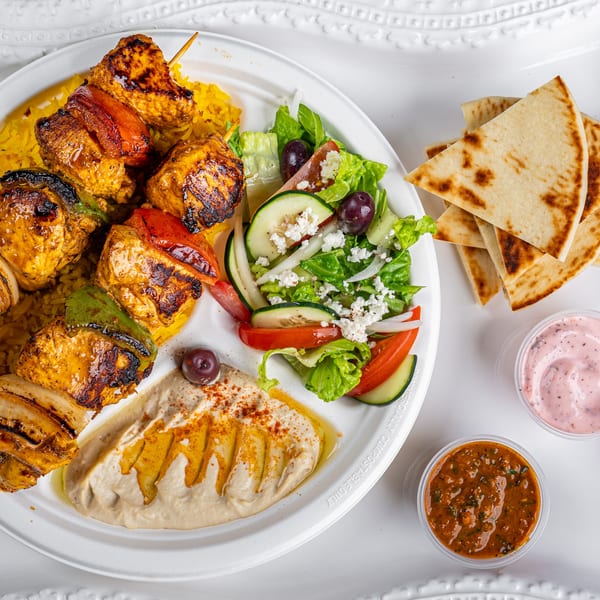 Greek chicken souvlaki plate with rice, salad, hummus, pita bread, and two dipping sauces.