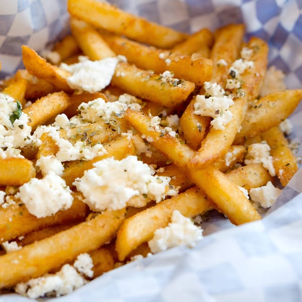 Golden french fries topped with crumbled feta cheese and herbs, served in a checkered paper container.