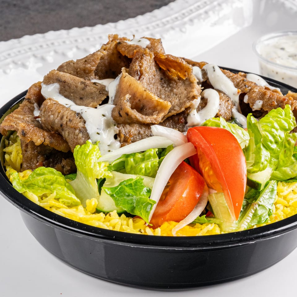 A Greek gyro bowl with sliced meat, rice, lettuce, tomatoes, onions, and tzatziki sauce.