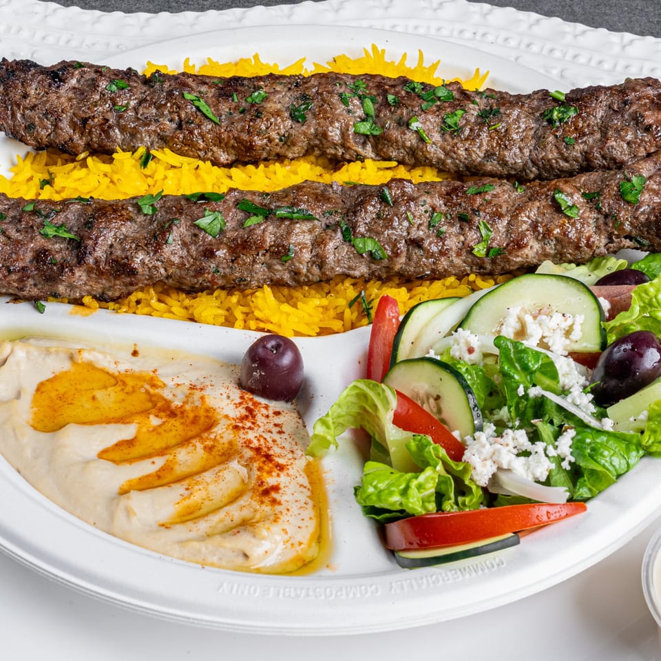 Two Greek beef kebabs on yellow rice, with hummus, salad, and an olive.