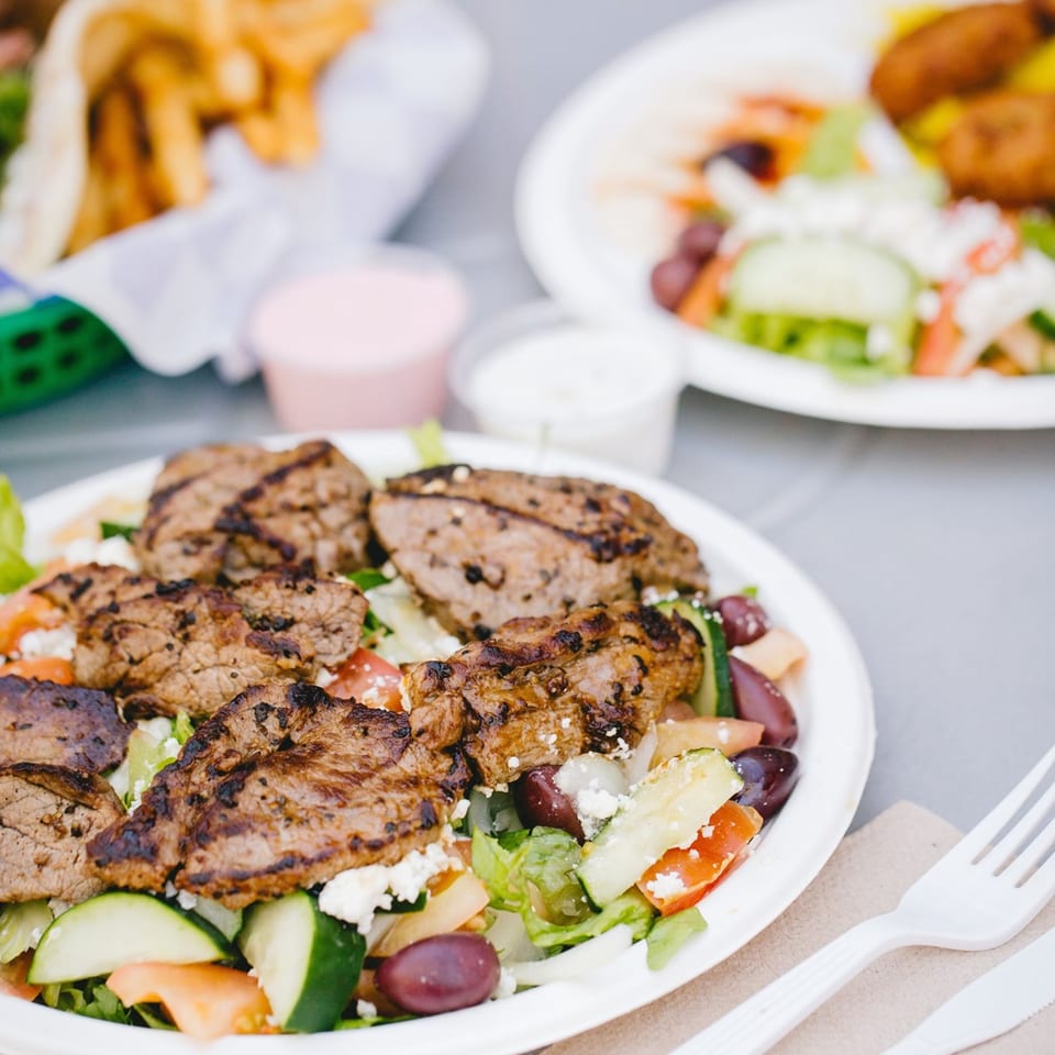 Grilled meat over a fresh Greek salad with feta, olives, and vegetables, with fries in the background.